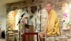 Master Mingtong Gu, dressed in a golden traditional Chinese jacket, sits smiling on a chair in a serene indoor space.